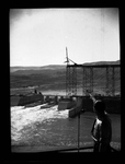 Man pointing to a crane working at the edge of a partially constructed railway bridge over the foundation of Grand Coulee Dam by Hubert Blonk