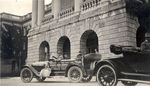 Official Mitchell Six touring car parked in front of the Great Lakes Naval Training Center with two men sitting inside it. by Frank W. Guilbert